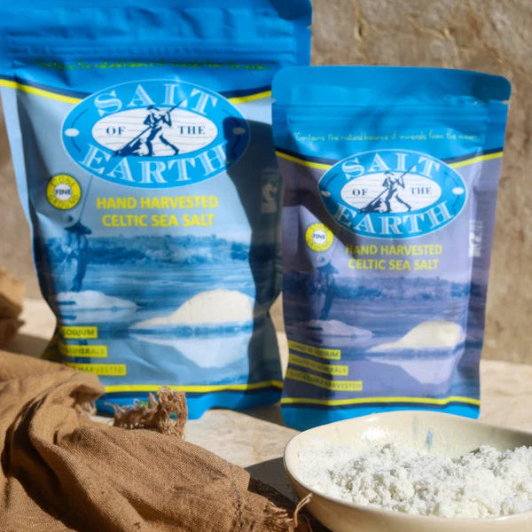 Two packages of 'Salt of the Earth' sea salt on a stone surface with a bowl of salt.